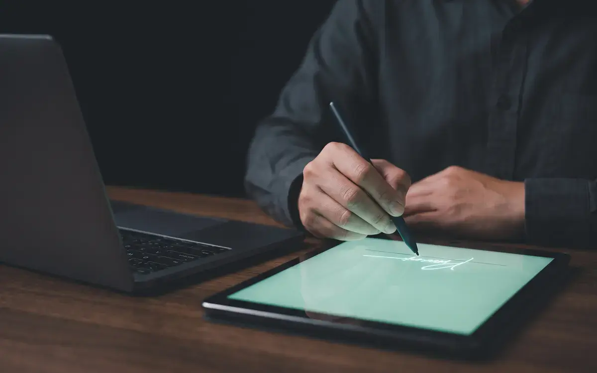 Someone signing a document on a tablet with a stylus, a laptop beside it.