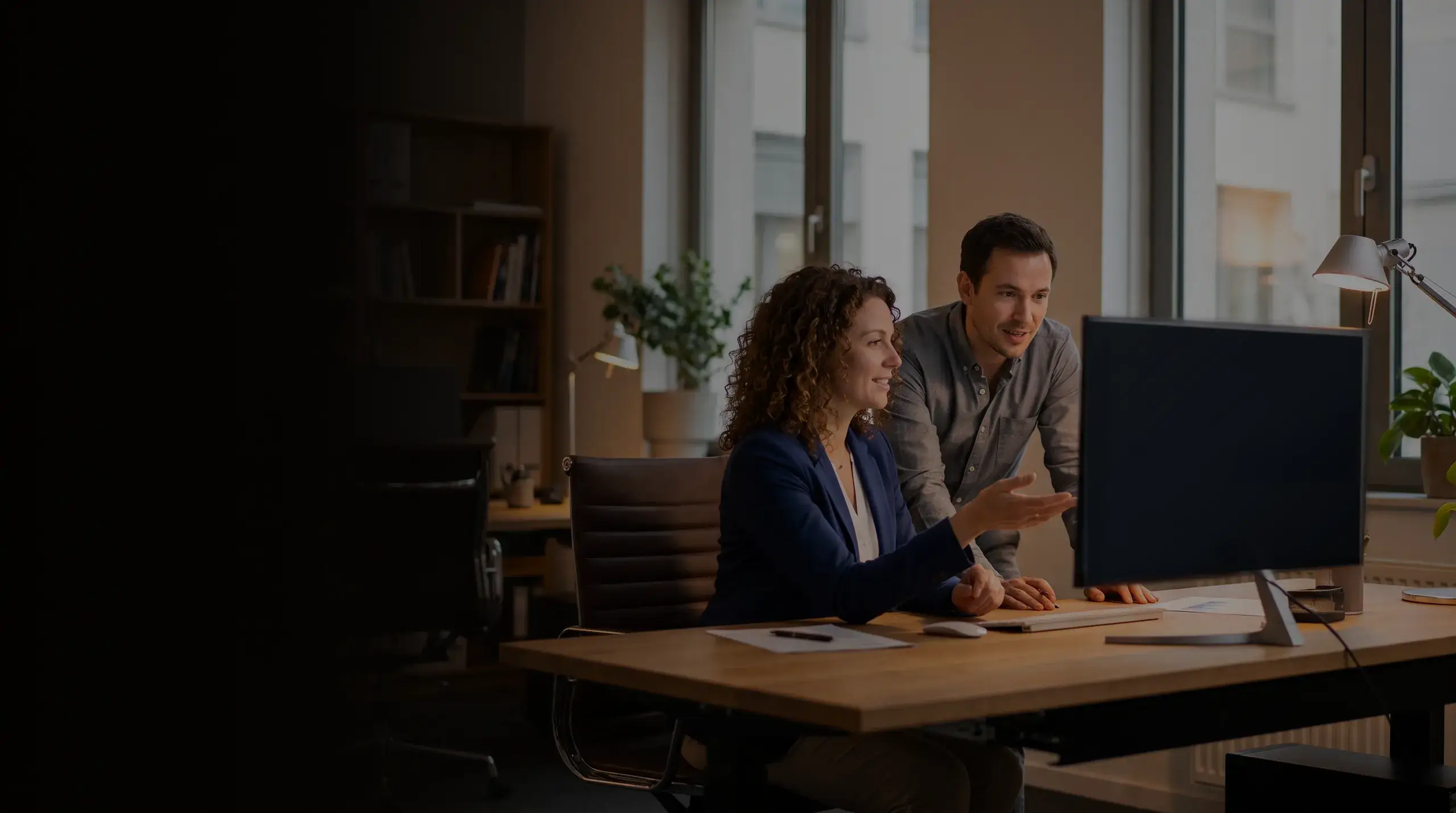 Two colleagues at a desk in a bright office, talking through work on a monitor.